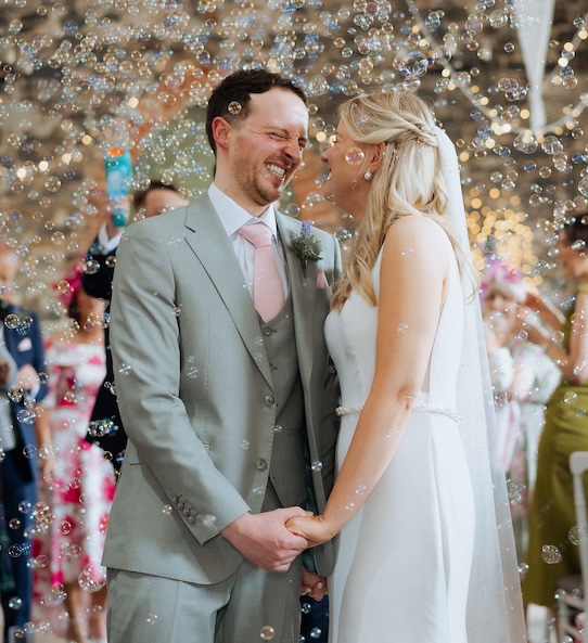 A newly married couple hold hands surrounded by their guests with bubbles in the air at Orange Tree House